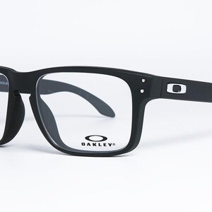 May include: Black square-framed spectacles with clear lenses. The glasses have a matte finish and the Oakley logo is visible on the frame. The temples are thick and curve down at the ends. The spectacles are set against a white background.