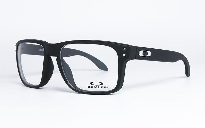 May include: Black square-framed spectacles with clear lenses. The glasses have a matte finish and the Oakley logo is visible on the frame. The temples are thick and curve down at the ends. The spectacles are set against a white background.