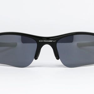 K&ouml;nnte beinhalten: Schwarze Sonnenbrille mit grauen Gl&auml;sern. Die Sonnenbrille hat einen geschwungenen Rahmen und einen Steg &uuml;ber der Nase. Der Markenname "Oakley" ist auf dem Rahmen sichtbar. Die Gl&auml;ser sind dunkelgrau.