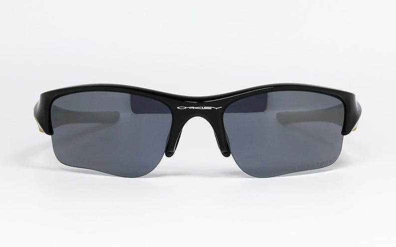 K&ouml;nnte beinhalten: Schwarze Sonnenbrille mit grauen Gl&auml;sern. Die Sonnenbrille hat einen geschwungenen Rahmen und einen Steg &uuml;ber der Nase. Der Markenname "Oakley" ist auf dem Rahmen sichtbar. Die Gl&auml;ser sind dunkelgrau.