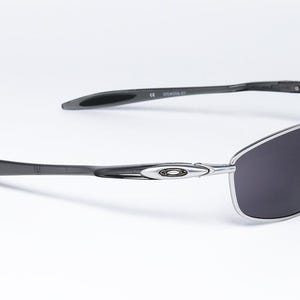 May include: Sleek, modern sunglasses with a silver frame and dark gray lenses. The temples are dark gray with a curved design. The Oakley logo is visible on the frame. The sunglasses are set against a white background.