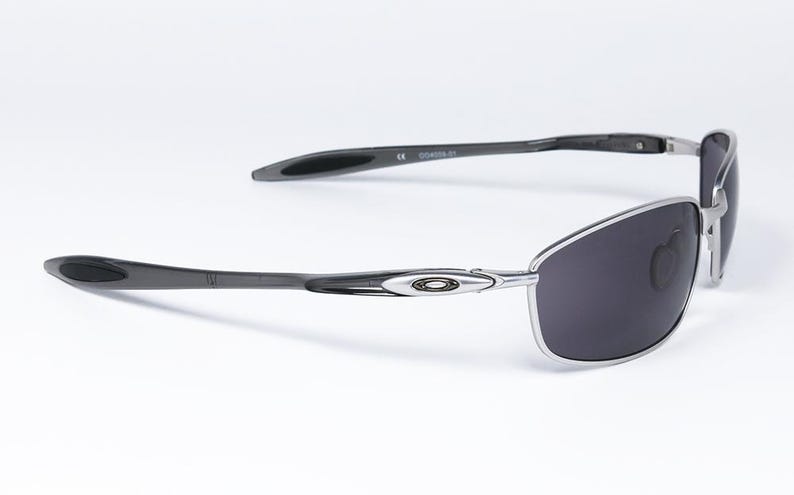 May include: Sleek, modern sunglasses with a silver frame and dark gray lenses. The temples are dark gray with a curved design. The Oakley logo is visible on the frame. The sunglasses are set against a white background.