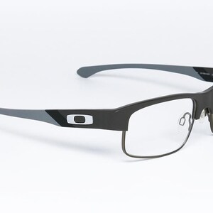 May include: Black and gray rectangular eyeglasses with clear lenses. The frames have a matte finish with a silver logo on the temples. The arms are gray and curve slightly. The glasses are set against a white background.