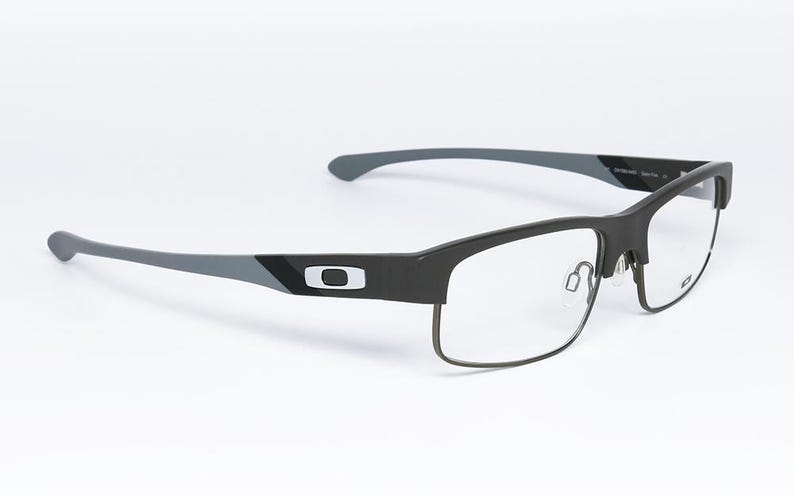 May include: Black and gray rectangular eyeglasses with clear lenses. The frames have a matte finish with a silver logo on the temples. The arms are gray and curve slightly. The glasses are set against a white background.