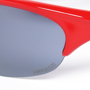 Puede incluir: Primer plano de gafas de sol rojas con lentes grises. Las gafas de sol tienen un dise&ntilde;o elegante y moderno con la palabra "DUCATI" impresa en la lente. La montura es de color rojo vibrante y las patillas son negras.