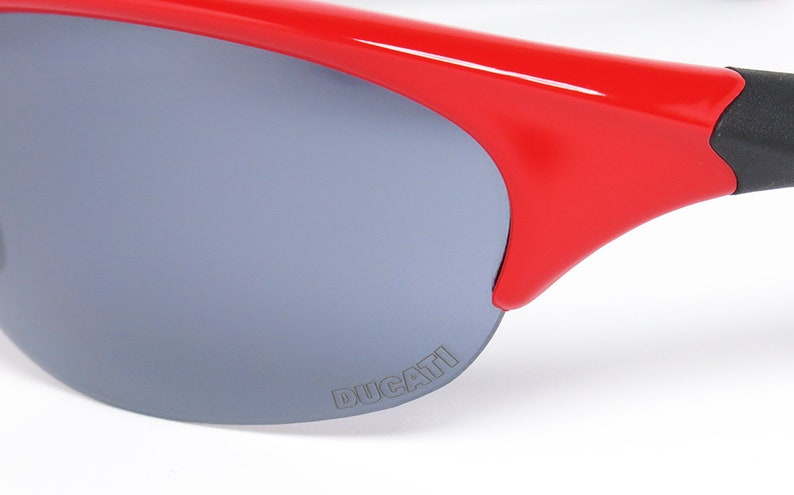 Puede incluir: Primer plano de gafas de sol rojas con lentes grises. Las gafas de sol tienen un dise&ntilde;o elegante y moderno con la palabra "DUCATI" impresa en la lente. La montura es de color rojo vibrante y las patillas son negras.
