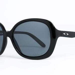 Puede incluir: Gafas de sol negras de gran tama&ntilde;o con lentes de color gris oscuro. Las gafas tienen una montura redondeada y un dise&ntilde;o elegante. El logotipo de Oakley es visible en las patillas. Las lentes est&aacute;n polarizadas.
