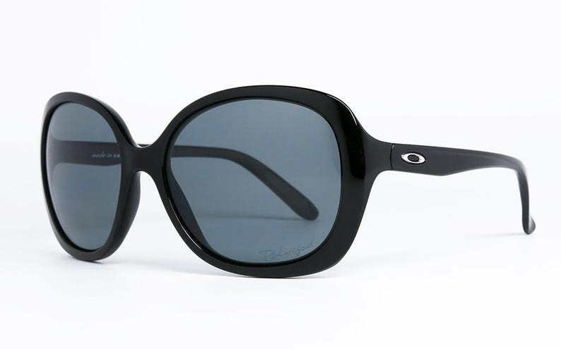Puede incluir: Gafas de sol negras de gran tama&ntilde;o con lentes de color gris oscuro. Las gafas tienen una montura redondeada y un dise&ntilde;o elegante. El logotipo de Oakley es visible en las patillas. Las lentes est&aacute;n polarizadas.