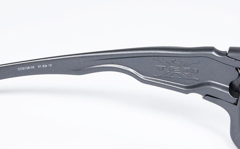 May include: Close-up of a gray sunglass arm with the word "TEN" embossed on the side. The arm also has the numbers "009128-08 61 16" printed on it. The sunglasses are likely for outdoor activities.