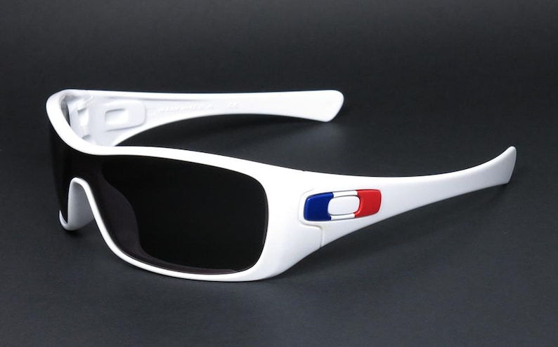 Op de afbeelding: Witte zonnebril met zwarte glazen. De bril heeft een wit montuur met een blauw-wit-rood embleem op de poot. Deze zonnebril is ontworpen voor sport en buitenactiviteiten.