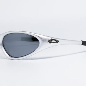 May include: Silver-framed sunglasses with dark lenses. The sunglasses have a sleek, curved design with a black accent on the temples. The Oakley logo is visible on the side. A modern eyewear accessory.