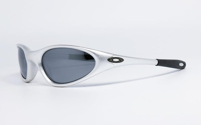 May include: Silver-framed sunglasses with dark lenses. The sunglasses have a sleek, curved design with a black accent on the temples. The Oakley logo is visible on the side. A modern eyewear accessory.
