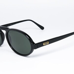 May include: Black oval sunglasses with dark lenses. The sunglasses have a bridge and arms, with a gold emblem on the temples. The frames are a glossy black, and the lenses are a dark green color.