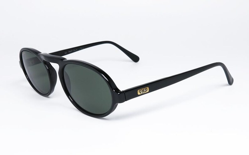 May include: Black oval sunglasses with dark lenses. The sunglasses have a bridge and arms, with a gold emblem on the temples. The frames are a glossy black, and the lenses are a dark green color.