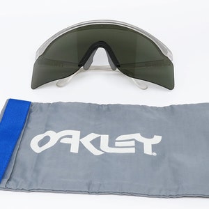 Puede incluir: Gafas de sol con montura transparente y lentes gris oscuro, con una funda de transporte de la marca Oakley gris con una correa azul.