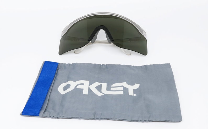 Puede incluir: Gafas de sol con montura transparente y lentes gris oscuro, con una funda de transporte de la marca Oakley gris con una correa azul.