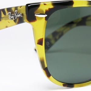 May include: Close-up of Ray-Ban sunglasses with a yellow and brown tortoise shell pattern. The sunglasses have dark green lenses and the Ray-Ban logo is visible on the temple. The frame is a classic Wayfarer style.