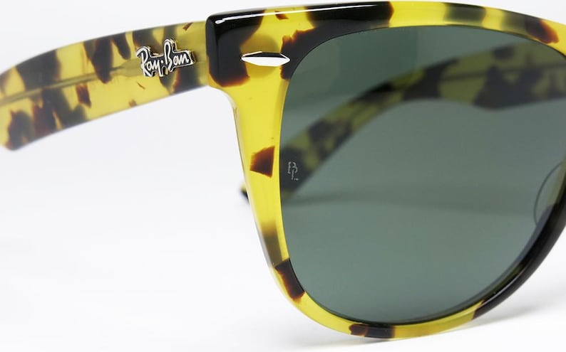May include: Close-up of Ray-Ban sunglasses with a yellow and brown tortoise shell pattern. The sunglasses have dark green lenses and the Ray-Ban logo is visible on the temple. The frame is a classic Wayfarer style.