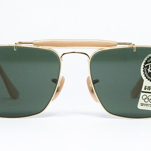 May include: Gold-framed Ray-Ban sunglasses with dark green lenses. The sunglasses have a bridge bar and a light-colored brow bar. A sticker on the lens reads "Ray-Ban" and "G-15 LENS". The sunglasses are a classic aviator style.