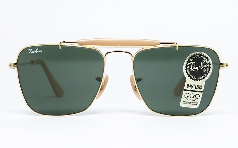 May include: Gold-framed Ray-Ban sunglasses with dark green lenses. The sunglasses have a bridge bar and a light-colored brow bar. A sticker on the lens reads "Ray-Ban" and "G-15 LENS". The sunglasses are a classic aviator style.