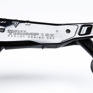 May include: Close-up of black Oakley sunglasses with the text "Oakley TESSERACT 1GB OAKLEY DESIGN USA" printed on the frame. The sunglasses have a sleek, modern design with a silver accent and a circular hinge.