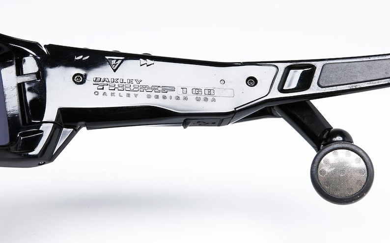 May include: Close-up of black Oakley sunglasses with the text "Oakley TESSERACT 1GB OAKLEY DESIGN USA" printed on the frame. The sunglasses have a sleek, modern design with a silver accent and a circular hinge.