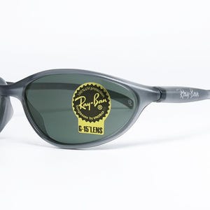 May include: Gray Ray-Ban sunglasses with dark lenses. The sunglasses have a wraparound design and a sticker that reads "Ray-Ban" and "G-15 LENS". The arms of the sunglasses are gray and have the Ray-Ban logo.