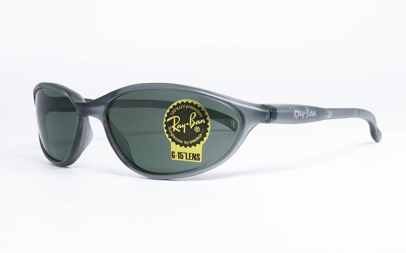 May include: Gray Ray-Ban sunglasses with dark lenses. The sunglasses have a wraparound design and a sticker that reads "Ray-Ban" and "G-15 LENS". The arms of the sunglasses are gray and have the Ray-Ban logo.