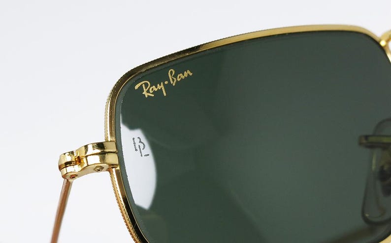 以下が含まれることがあります： ゴールドフレームと濃い緑色のレンズを備えたRay-Banサングラスのクローズアップ。 Ray-Banのロゴは、左上のレンズに金色の文字で表示されています。 ヒンジとテンプルアームも金色です。