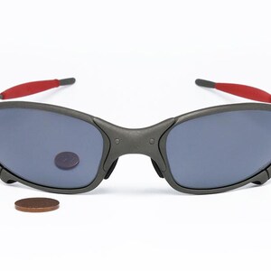 May include: Dark gray sunglasses with blue lenses and red accents on the temples. The sunglasses are displayed on a white background. A penny is next to the sunglasses.