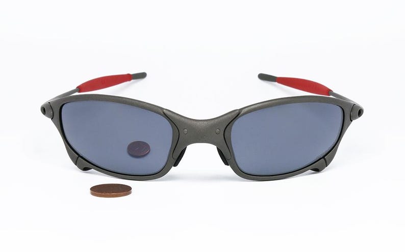 May include: Dark gray sunglasses with blue lenses and red accents on the temples. The sunglasses are displayed on a white background. A penny is next to the sunglasses.