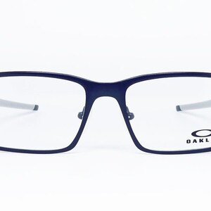 K&ouml;nnte beinhalten: Rechteckige Brille in Marineblau mit silbernen B&uuml;geln. Die Brille hat einen dunkelblauen Rahmen und klare Gl&auml;ser. Das Oakley-Logo ist auf dem rechten Glas zu sehen. Die Brille ist vor wei&szlig;em Hintergrund platziert.