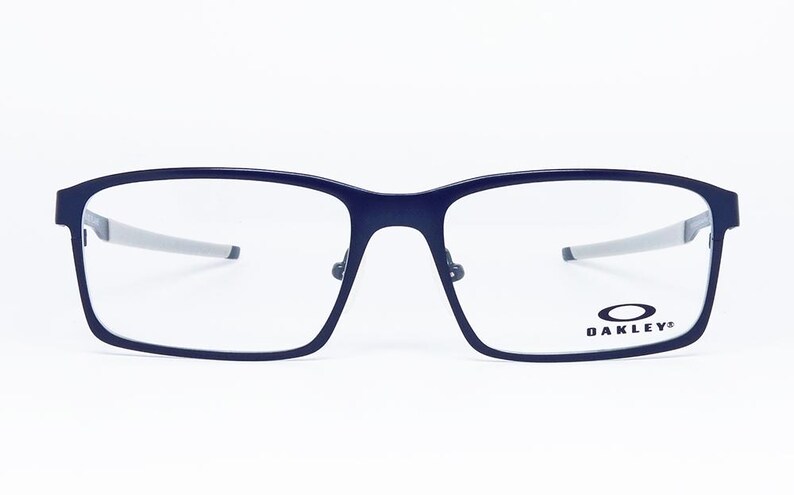 K&ouml;nnte beinhalten: Rechteckige Brille in Marineblau mit silbernen B&uuml;geln. Die Brille hat einen dunkelblauen Rahmen und klare Gl&auml;ser. Das Oakley-Logo ist auf dem rechten Glas zu sehen. Die Brille ist vor wei&szlig;em Hintergrund platziert.