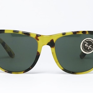 May include: Sunglasses with a classic design. The frames are a yellow and brown tortoiseshell pattern, with dark lenses. A round sticker with the Ray-Ban logo is on the right lens. The sunglasses are set against a white background.