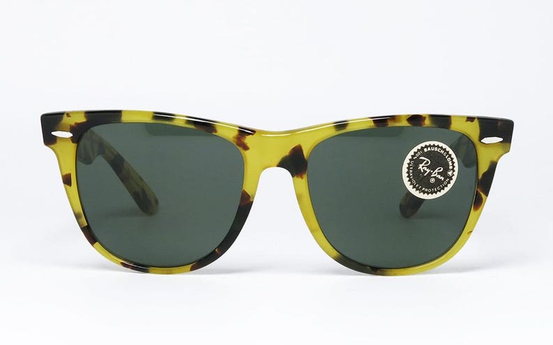 May include: Sunglasses with a classic design. The frames are a yellow and brown tortoiseshell pattern, with dark lenses. A round sticker with the Ray-Ban logo is on the right lens. The sunglasses are set against a white background.