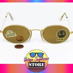 Pu&ograve; includere: Occhiali da sole Ray-Ban B-15 con montatura dorata e lenti ovali. Gli occhiali da sole hanno un ponte e aste dorate e sono esposti con una moneta e un adesivo. L'immagine include il logo di Vintage Sunglasses Store.
