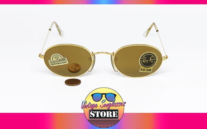 Pu&ograve; includere: Occhiali da sole Ray-Ban B-15 con montatura dorata e lenti ovali. Gli occhiali da sole hanno un ponte e aste dorate e sono esposti con una moneta e un adesivo. L'immagine include il logo di Vintage Sunglasses Store.