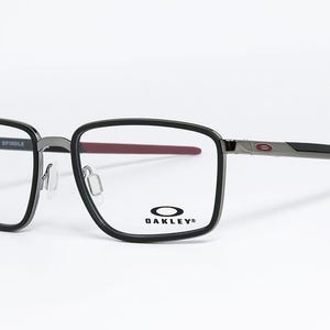 May include: Black and gray Oakley Spindle eyeglasses with a rectangular frame. The temples are gray and black with a red accent at the end. The Oakley logo is visible on the lens.