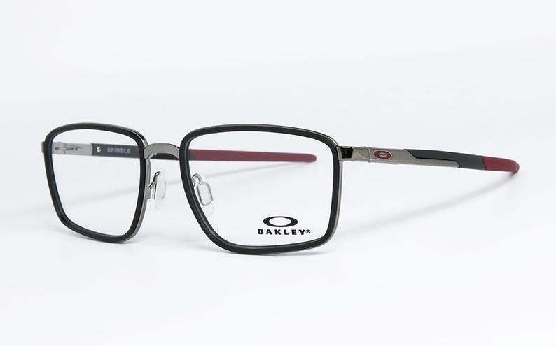 May include: Black and gray Oakley Spindle eyeglasses with a rectangular frame. The temples are gray and black with a red accent at the end. The Oakley logo is visible on the lens.