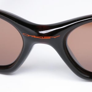 Pode incluir: Close-up de &oacute;culos de sol com arma&ccedil;&atilde;o castanha escura e lentes castanhas. O nome da marca "Oakley" est&aacute; impresso em laranja na ponte. Os &oacute;culos de sol t&ecirc;m um design curvo.