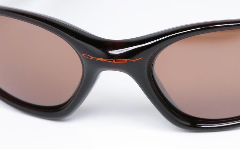 Pode incluir: Close-up de &oacute;culos de sol com arma&ccedil;&atilde;o castanha escura e lentes castanhas. O nome da marca "Oakley" est&aacute; impresso em laranja na ponte. Os &oacute;culos de sol t&ecirc;m um design curvo.