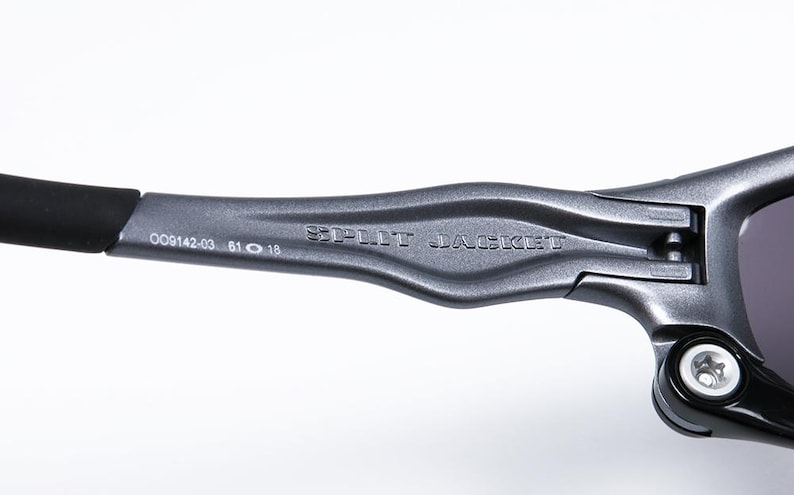 Puede incluir: Primer plano de una montura de gafas de sol de metal gris con el texto "SPLIT JACKET" grabado en el lateral. La montura tiene una punta de varilla de goma negra y el texto "OO9142-03 61 0 18" est&aacute; impreso en el lateral.
