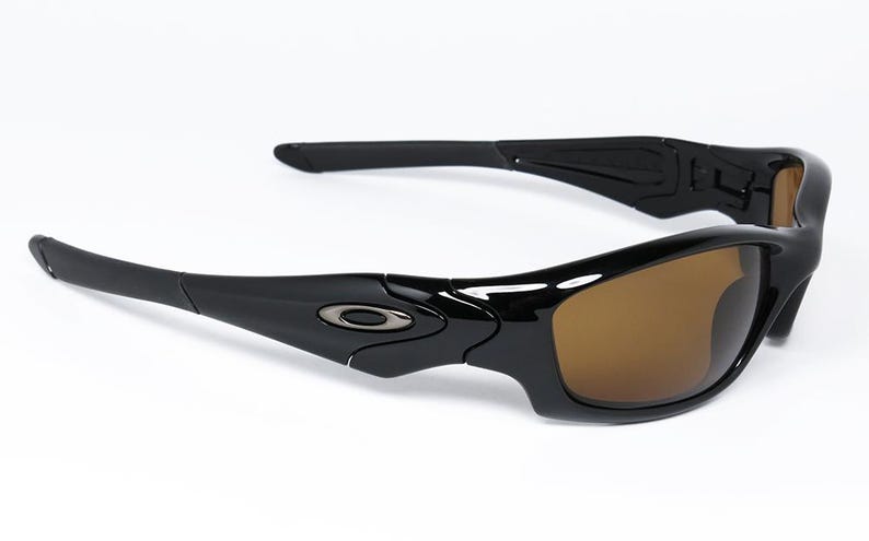 May include: Black sunglasses with brown lenses. The sunglasses have a sleek, modern design with a glossy finish. The Oakley logo is visible on the temples. Suitable for sports and outdoor activities.