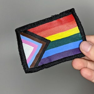 May include: Rectangular patch featuring the Progress Pride flag design. The patch has a black border and includes the traditional rainbow colors, along with additional colors representing transgender, Black, and marginalized communities.