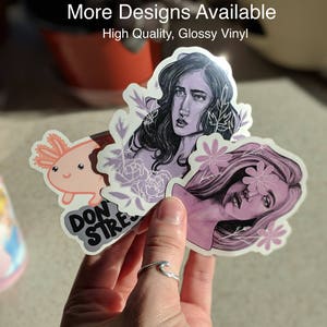 May include: A hand holding a collection of glossy vinyl stickers. The stickers feature illustrations of a woman, an axolotl, and the words "DON'T STRESS". The text "More Designs Available" and "High Quality, Glossy Vinyl" are also visible.