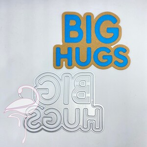 Puede incluir: Una troqueladora de metal para proyectos de manualidades con las palabras "BIG HUGS" en una fuente en negrita y con contorno. La troqueladora es plateada y tiene un acabado metálico liso.