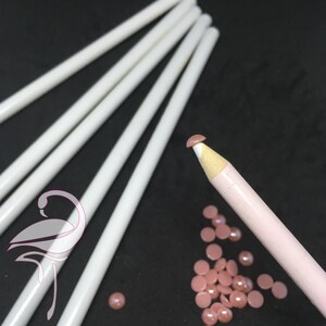 Puede incluir: Primer plano de un lápiz rosa y blanco con punta redondeada. El lápiz está rodeado de pequeñas piedras de cristal redondas de color rosa. También hay varios lápices blancos en el fondo.