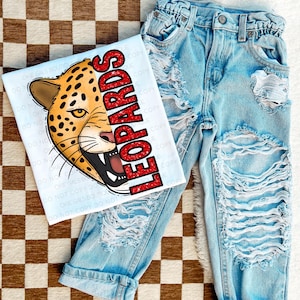 May include: A white t-shirt with a red glitter graphic of a leopard's face and the word "LEOPARDS" on it. The shirt is paired with light blue distressed denim jeans.