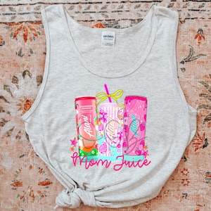 Pu&ograve; includere: Canotta grigio chiaro con una grafica colorata di tre bevande. I contenitori sono rosa e bianchi, con il testo "Alani" e "Mom Juice" in rosa. Il design include stelle e frutta.