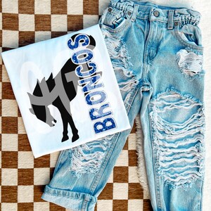 Può includere: Una maglietta bianca con una grafica blu glitterata che dice "Broncos" e una silhouette nera e grigia di un cavallo. La maglietta è abbinata a jeans di denim blu chiaro con dettagli usurati.